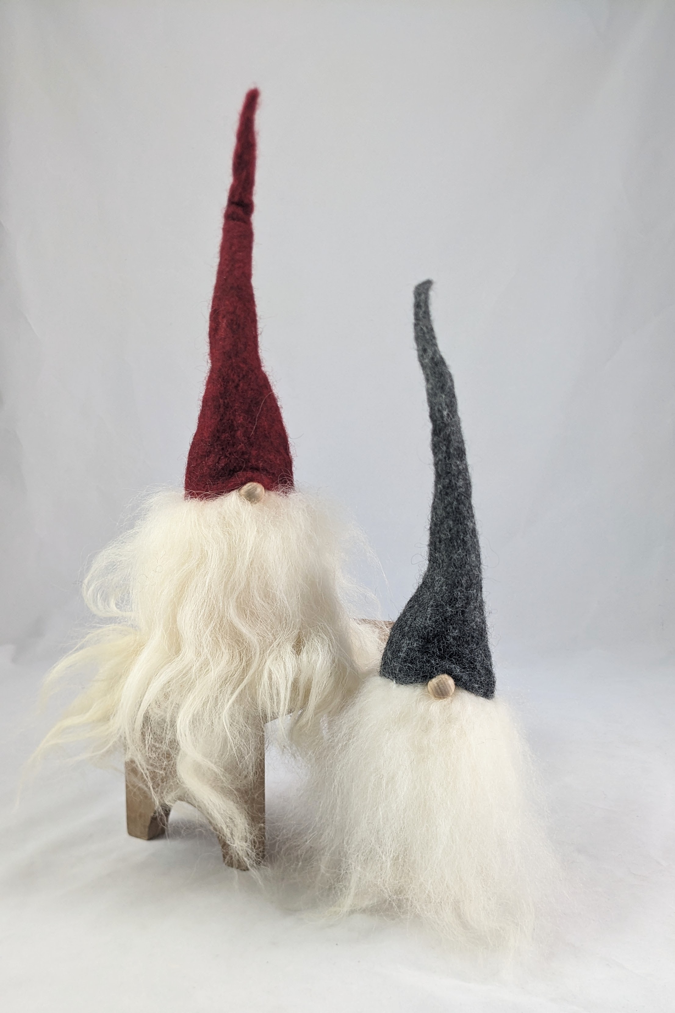 2 tomtes with long wool beards, one with a burgundy tall hat is propped up on a wooden stand and the base is level with the nose of the gray hat tomte on the ground.