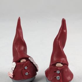 Two kid tomtes, one with only a nose showing and the other two braids and a nose. Both are dressed in red overcoats although there are no arms, and a red tall hat. 