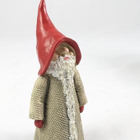 Santa Martin standing at an angle, with a bright red pointed hat and red gloves. The narrow beard goes all the way to the floor covering the front of a textured beige overcoat.
