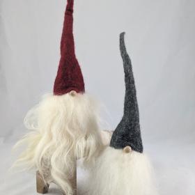 2 tomtes with long wool beards, one with a burgundy tall hat is propped up on a wooden stand and the base is level with the nose of the gray hat tomte on the ground.