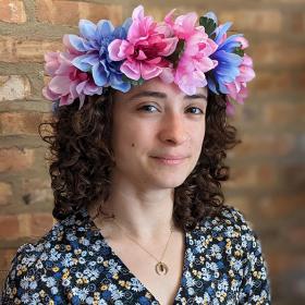 Photo portrait of a woman with curly brown hair, wearing a crown of pink and purple flowers, in front of a brick wall